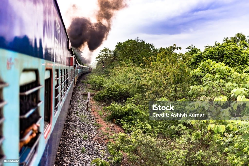 Beautiful photo of an Indian Railways Train drawn by diesel engine, and emitting smoke while passing through a lush green forest area in the rainy season. Photo depicting that Journey is more beautiful than the Destination. The photo is taken in rainy season of monsoons while travelling in the iconic Goa Express of Indian Railways being ran on Diesel. Due to tough terrains the route is yet to be electrified. Air Pollution Stock Photo