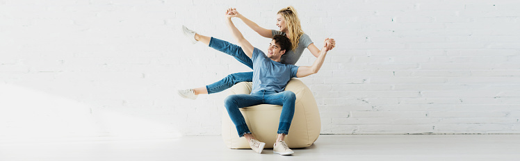 panoramic shot of cheerful blonde girl and happy man holding hands while sitting on bean bag chair