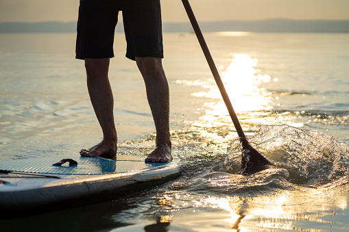 SUP detail legs of man standing on stand up paddle board paddling in lake at sunset summer reflections in water