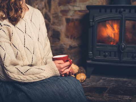 A young woman in a white jumper is sitting by the fireplace drinking from a mug