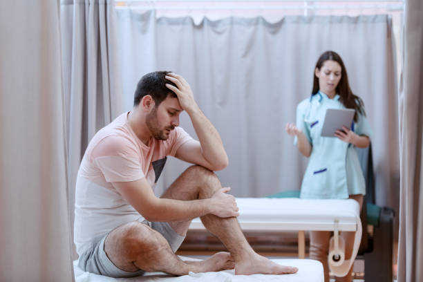 giovane paziente maschio caucasico seduto sul letto d'ospedale e che prova dolore. in background infermiera femminile usando tablet. - emergency room accident hospital emergency sign foto e immagini stock