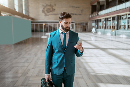 Young attractive bearded serious businessman in blue suit carrying luggage and using smart phone while standing at train station. Business trip concept.