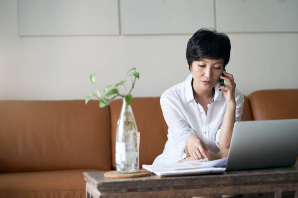 donna professionista asiatica che parla allo smartphone mentre lavora a casa - laptop adult curtain business foto e immagini stock