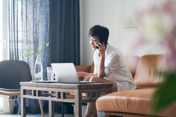 asiatische selbstständige im gespräch mit smartphone während der arbeit am laptop zu hause - laptop adult curtain business stock-fotos und bilder