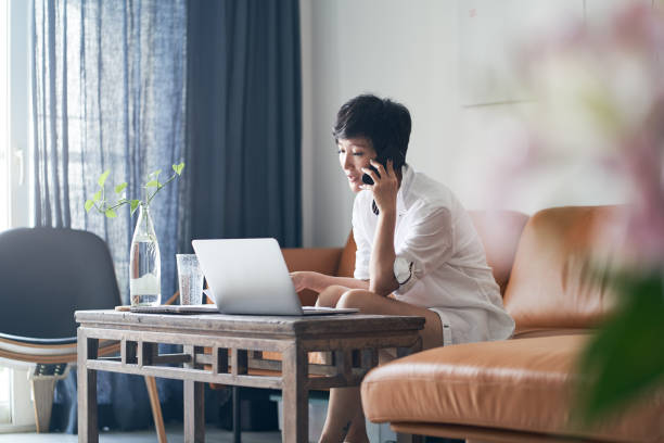 Asian self employed woman talking on smart phone while working on laptop at home stock photo