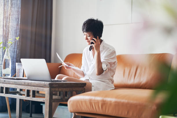 Asian female freelancer talking on smart phone while working on laptop at home stock photo