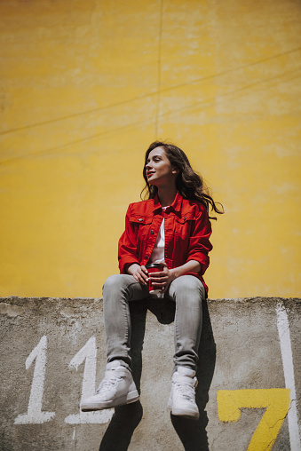 Urban lifestyle concept. Low angle full length portrait of young smiling hipster lady holding cup of coffee while sitting on parapet and looking far away