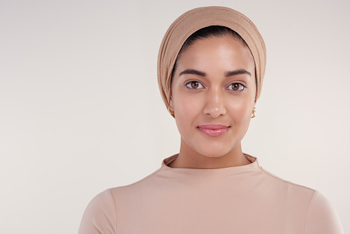 Studio shot of an attractive young woman wearing a headscarf smiling at the camera against a beige background
