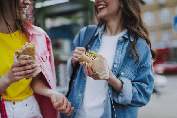 zwei glückliche freundinnen, die hot dogs essen, essen sie zu fuß - people eating walking fun stock-fotos und bilder