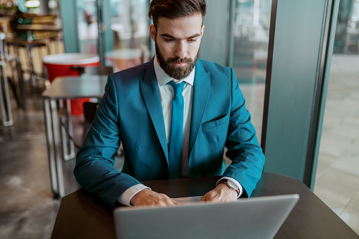Young adult focused businessman in blue suit sitting at cafe and using laptop for work.
