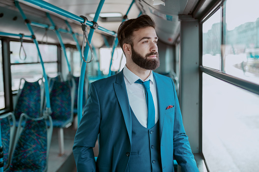 Young thoughtful bearded businessman in blue suit standing in public transportation and looking trough window.