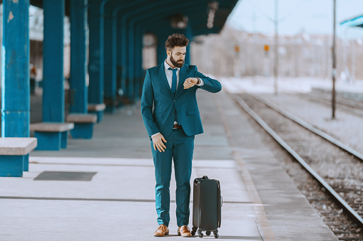 Young handsome bearded businessman in blue suit looking at wristwatch and waiting for train to come. Next to him is luggage. Business trip concept.