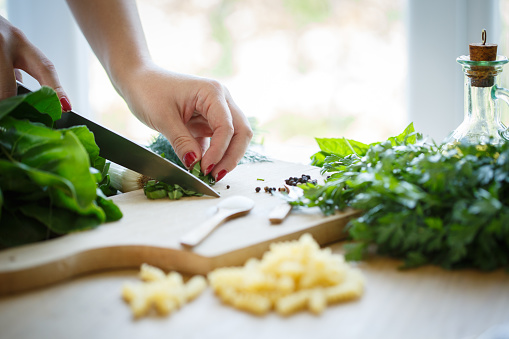 Close up shot of unrecognizable female hand cutting up fresh basil on a cutting board surrounded with fresh herbs and pasta.