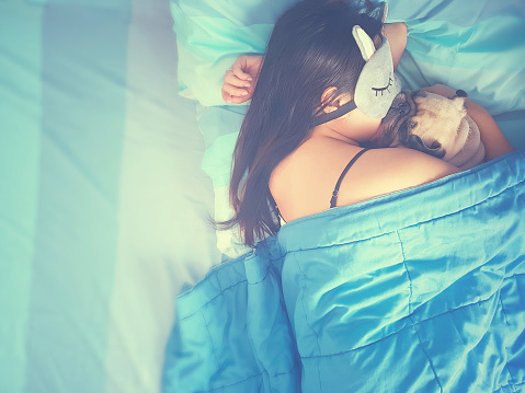 Young woman is lying and sleeping with pug dog puppy in the bed. Nap with hugging dog