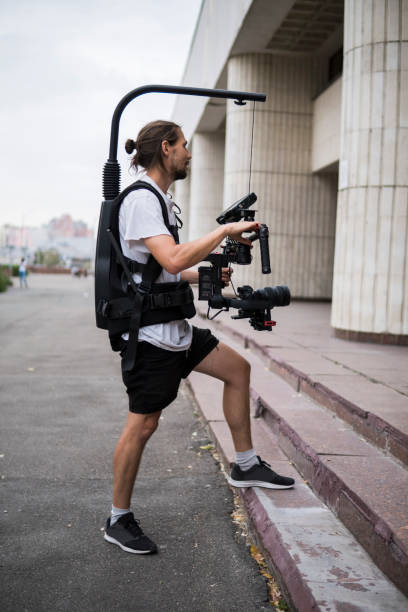 istock Professional videographer holding camera on 3-axis gimbal which mounted on easy rig. Videographer using steadicam. Pro equipment helps to make high quality video without shaking. 1151427161