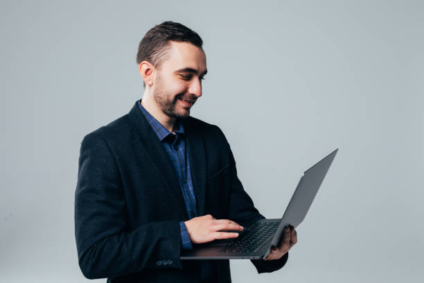istock Side view of attractive young businessman in classical suit using laptop, standing against gray background 1151436619