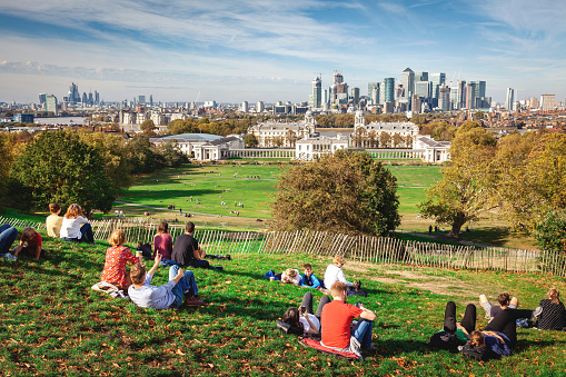London, United Kingdom - October 13, 2018: Relaxing in Greenwich Park overlooking London. Young people relax in Greenwich Park on a summer afternoon and await the sunset. Greenwich Park is the oldest enclosed Royal Park and is situated on a hilltop, it has great views over London including the Naval College, Canary Wharf, the Millennium Dome and the skyscrapers of the city of London.