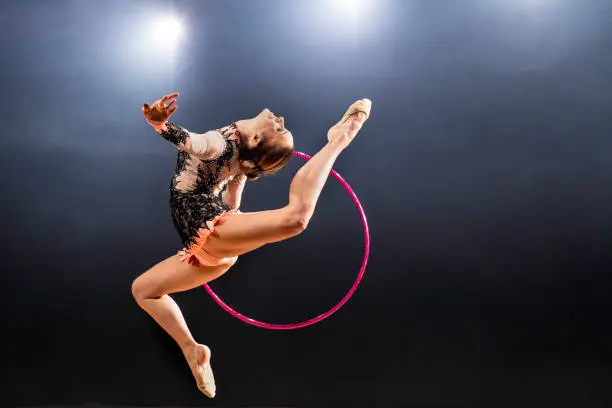 Studio shot of a young beautiful rhytmic gymnast with gymnastic ring doing mid air jump in front of a black background,  copy space.