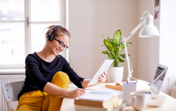 A young female student sitting at the table, using tablet when studying. A young happy college female student sitting at the table at home, using tablet and headphones when studying. music theory stock pictures, royalty-free photos & images