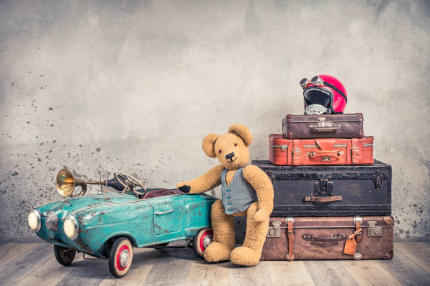 teddy bear toy standing near rusty retro  pedal car from 60s, antique travel trunks luggage, old leather valises, red helmet with outdated goggles front loft background. vintage style filtered photo - trunk luggage old fashioned retro revival imagens e fotografias de stock