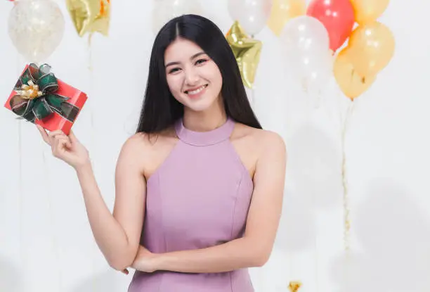 Beautiful young asian woman big friendly smile posig with gift box happy at fun party, portrait shot white background with colorful balloons.
