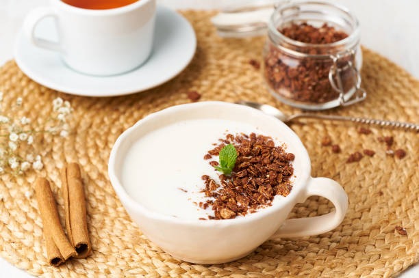 Yogurt with chocolate granola in cup, breakfast with tea on beige background, side view.