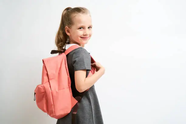 Photo of Little girl photographed against white background wearing school uniform dress isolated holding a coral backpack on both shoulders