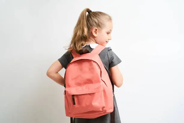 Photo of Little girl photographed against white background wearing school uniform dress isolated holding a coral backpack on both shoulders