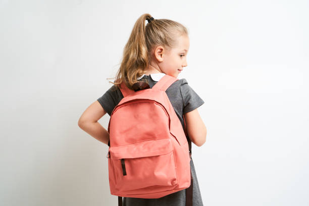 little girl photographed against white background wearing school uniform dress isolated holding a coral backpack on both shoulders - mochila imagens e fotografias de stock