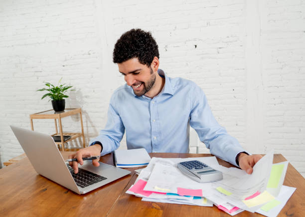 felice imprenditore attraente che calcola costi, spese, mutui, tasse o paga bollette con documenti e laptop all'ufficio di casa. nell'online banking e nelle finanze aziendali di successo libere da debiti. - mutuo proprietario foto e immagini stock