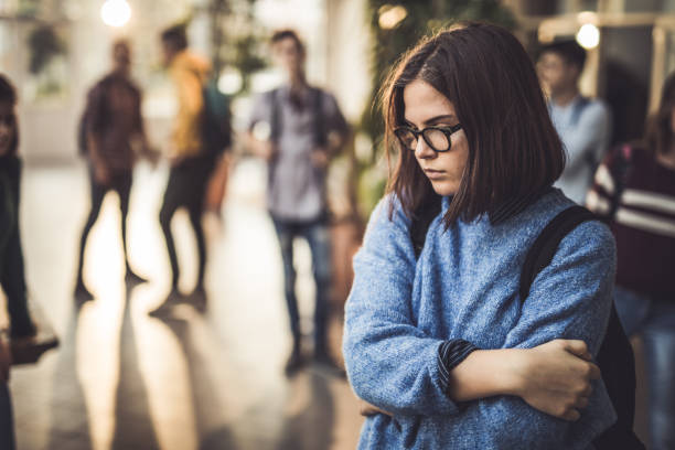 Sad high school student feeling lonely in a hallway. Displeased female student bullied by her classmate standing alone in a hallway. teenager stock pictures, royalty-free photos & images