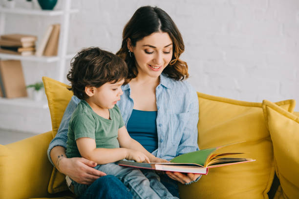 mama und sohn sitzen auf gelbem sofa und lesebuch - familie mit einem kind stock-fotos und bilder