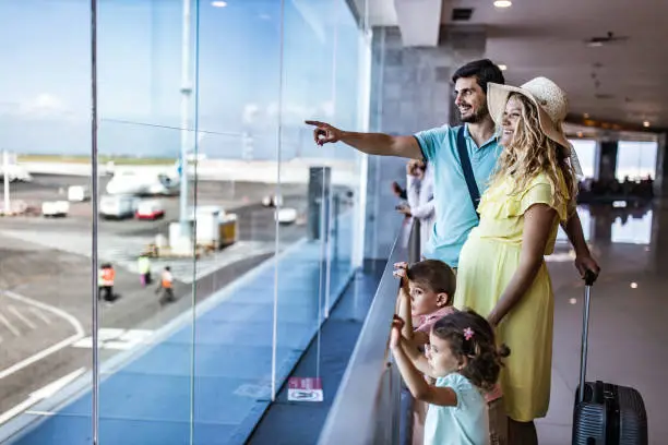 Happy family standing at the airport terminal and looking through window while man is aiming at distance. Woman is pregnant.