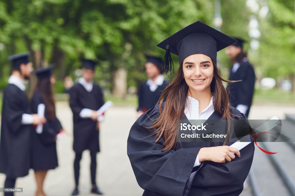 A young female graduate against the background of university graduates. A young female graduate with a scroll in her hands is smiling against the background of university graduates. Graduation.University gesture and people concept. Graduation Stock Photo
