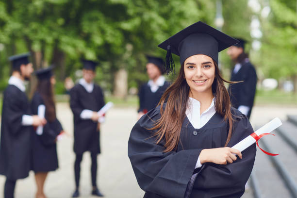 A young female graduate against the background of university graduates. A young female graduate with a scroll in her hands is smiling against the background of university graduates. Graduation.University gesture and people concept. College Graduates stock pictures, royalty-free photos & images