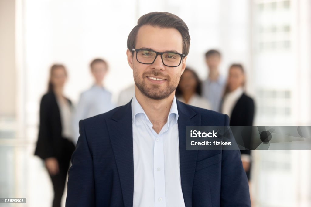 Confident businessman manager looking at camera with team on background Confident business man coach in suit director company owner professional manager looking at camera with team on background, smiling male ceo leader banker employer posing in office, headshot portrait Businessman Stock Photo