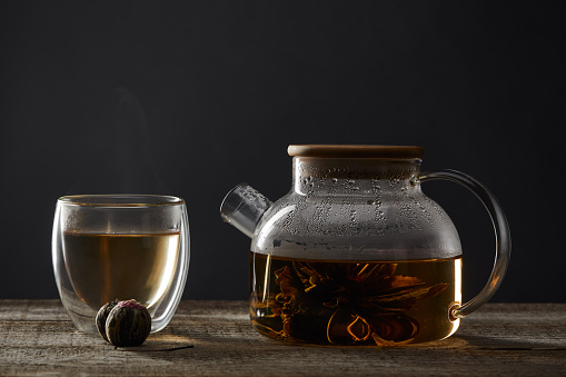 transparent teapot and glass with blooming tea on wooden table isolated on black