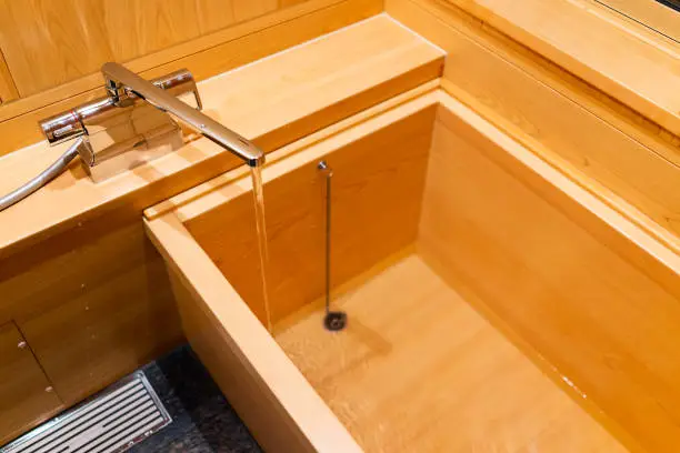 Photo of New clean cypress bathtub wooden traditional Japanese tub in home house or onsen hotel bathroom interior with nobody and running water in Japan