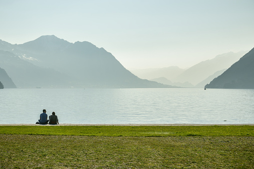 Brunnen, Switzerland - March 30, 2019: Young couple sitting on shores of Lake Luzern in small park in Brunnen, Switzerland during first warm day in spring 2019