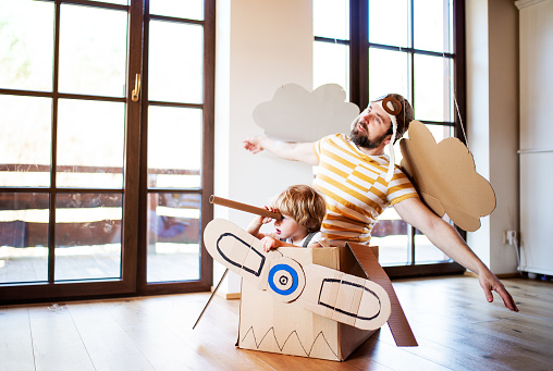 A toddler boy and father with carton plane playing indoors at home, flying concept.