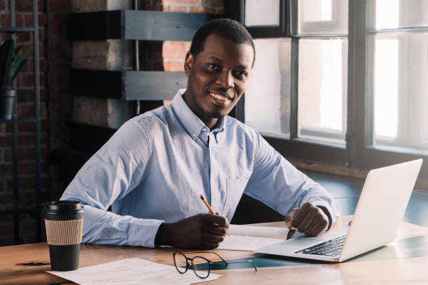 istock Young African male writing in papers while and using laptop during break in cafe 1151641789