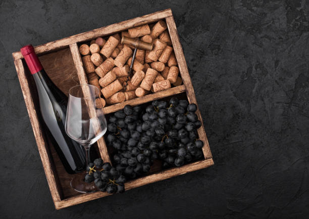 istock Bottle of red wine and empty glass with dark grapes with corks and corkscrew inside vintage wooden box on black stone background. Top view 1151667452