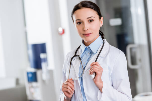 istock Young doctor in white coat touching stethoscope and looking at camera 1151675519