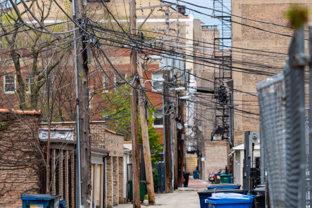 Alley with power lines and chain link fence An alleyway in daylight. Utility poles and power lines cross the sky. A bent chain link, dumpsters, garbage bins and a fire escape line the alley. seedy alley stock pictures, royalty-free photos & images