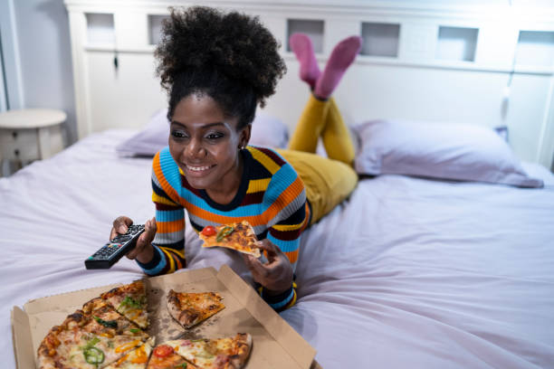 African woman lying down on bed and eating pizza