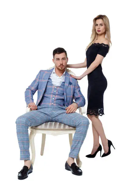 Photo of Front view of a luxury couple. A man is sitting on an expensive chair, a woman in a dress is standing next to him.