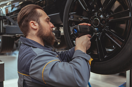 Bearded male car mechanic torquing the lug nuts of a wheel of a lifted car at his garage. Experienced automobile technician repairing an auto at his workshop