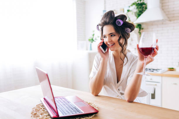 junge frau steht in küche und telefoniert rotweinglas in der hand halten. hausmeister mit curler im haar. hausfrau sorgloses leben. - stereotypical housewife women domestic kitchen brown hair stock-fotos und bilder