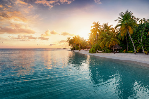 Sunrise behind a tropical island with palm trees, sandy beach and emerald sea in the Maldives