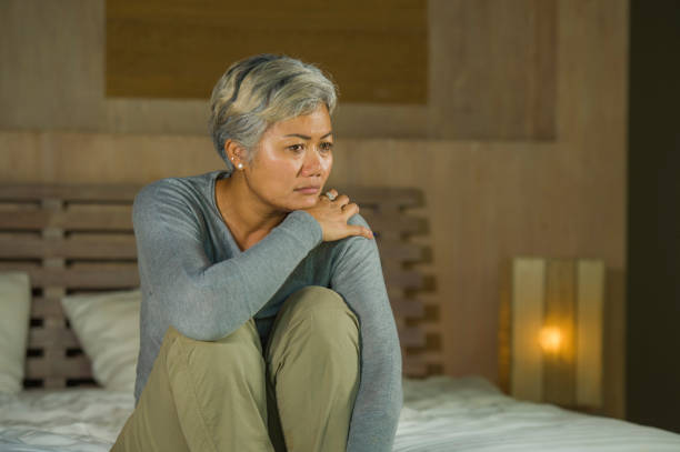 depressed and sad grey hair mature woman crying lonely sitting on bed suffering crisis in pain and depression problem feeling lost and overwhelmed at home bedroom frustrated and stressed stock photo